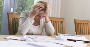A woman sits at the table, hands on her head, stressed over her bills
